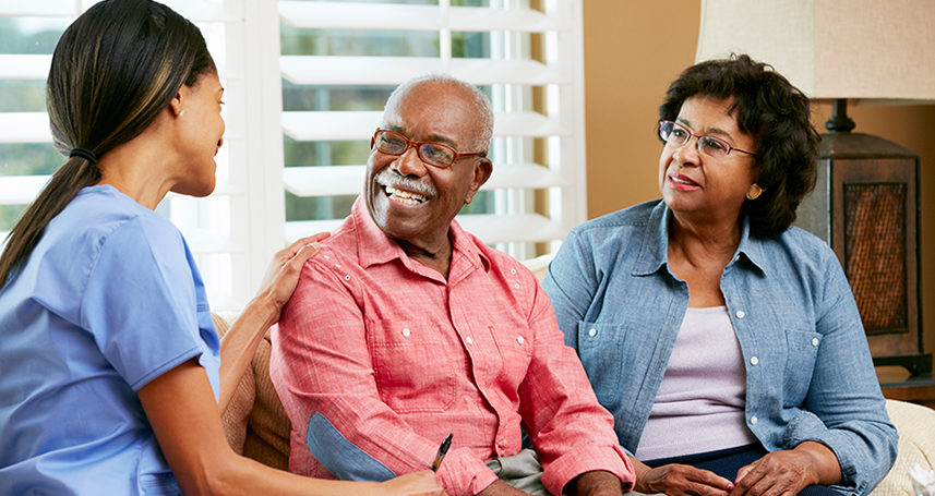 Nurse Making Notes During Home Visit With Senior Couple Smiling