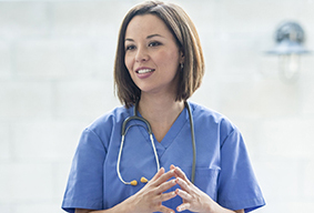 A multi-ethnic group of medical staff are indoors in a hospital. They are wearing medical clothing. A Caucasian female doctor is giving a presentation to the others.